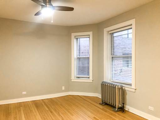 A room with a ceiling fan, two windows with blinds, and a radiator on the floor.