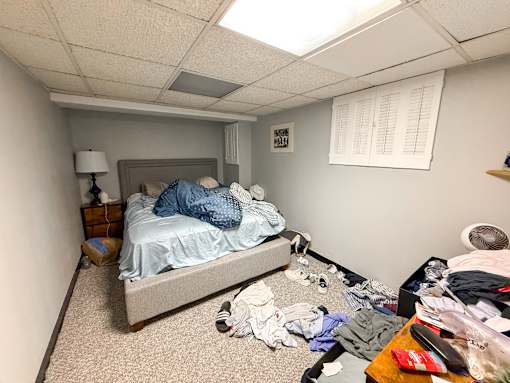 A messy bedroom with a bed, dresser, and clothes scattered on the floor.