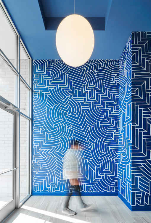 A person in a white sweater is walking through a room with a blue maze pattern on the wall.