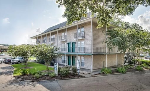 A building with a balcony and a green door is surrounded by trees and has a parking lot in front.