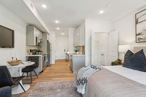 A modern bedroom with a large bed and a kitchen area in the background.