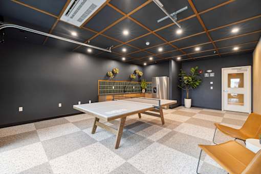 A room with a ping pong table, chairs and a fridge.