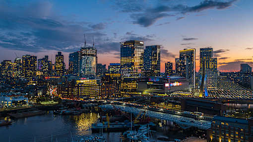 A city skyline at dusk with buildings illuminated and a bridge in the foreground.