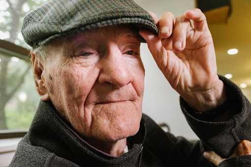 An elderly man wearing a green cap and black coat.