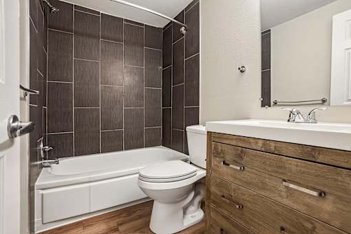 A white toilet sits next to a bathtub in a bathroom with wooden floors and dark tiled walls.