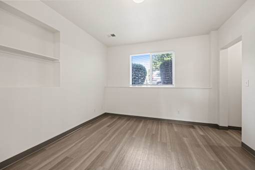 A room with wooden flooring and a window showing a view of trees and a building outside.