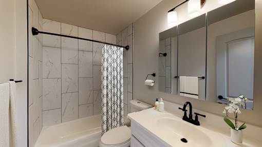 A bathroom with a white sink, a white toilet, a white towel, a white vase with white flowers, a white shower curtain with a black and white pattern, a white bathtub, a white towel