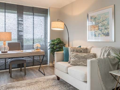 A living room with a white couch, a desk with a chair, a floor lamp, and a framed picture on the wall.