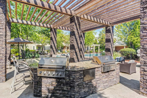 Townhomes In Aurora, IL - The Apartments At Kirkland Crossing - Covered Grill Area With Two BBQs, Trees, Counterspace With Bar Chairs.