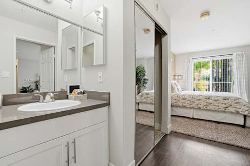 A bathroom with a sink, mirror, and a bedroom with a bed and window.