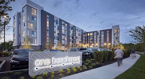 A modern apartment complex named One Beachmont is shown with a lit sign in front.