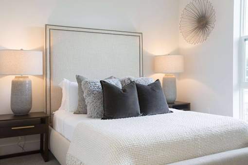 A bed with a white comforter and grey pillows is flanked by two lamps on a nightstand.