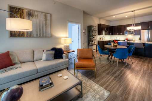 A modern living room with a grey sofa, a wooden coffee table, and a dining area with blue chairs.