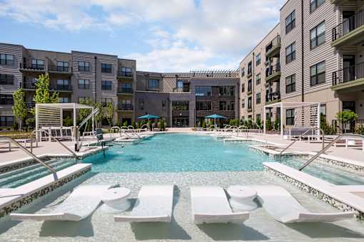 A large swimming pool surrounded by white lounge chairs and buildings in the background.