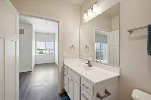 A bright bathroom with a white vanity, large mirror, and modern light fixtures. The room features a towel bar and a glimpse of the adjacent room visible through the door.
