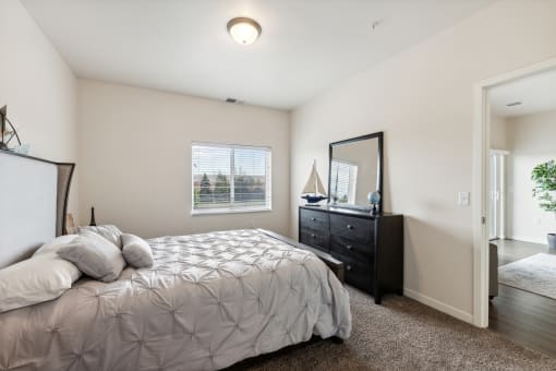 A cozy bedroom with a bed, decorative pillows, a dresser, and a mirror. The room features a window with natural light and leads into another space through an open door.