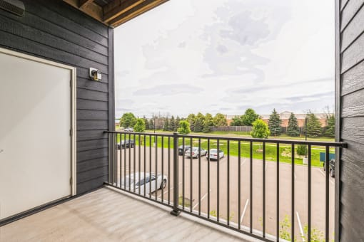 A balcony with a metal railing, overlooking a parking lot and green trees in the distance. The view is framed by the building's exterior wall and a door.
