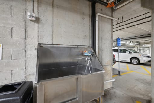 A stainless steel utility sink located in a parking garage, with a hose and a trash can nearby. A white vehicle is visible in the background.