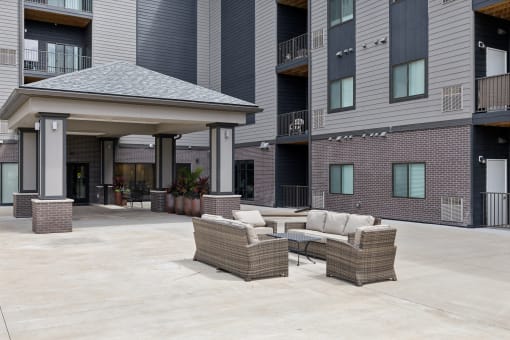 An outdoor seating area with a wicker sofa set, including chairs and a coffee table, located under a covered pavilion. The space is surrounded by modern apartment buildings.
