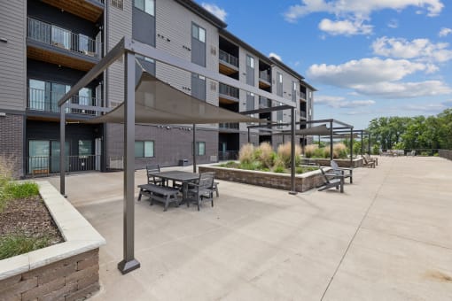 An outdoor seating area with shaded tables under canopies, surrounded by landscaped planters and modern apartment buildings. The area features a paved pathway and clear blue skies in the background.