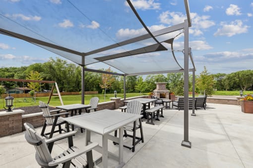 An outdoor patio area with shaded seating, including tables and chairs, under a canopy. The space features a view of green trees and a swing set in the distance.
