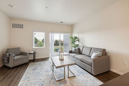 A cozy living room featuring a sofa, a coffee table, a decorative plant, and large windows letting in natural light. There's also a sliding glass door that opens to an outdoor area.