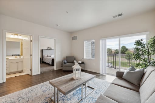 A bright living room with a sofa, coffee table, and decorative items. The space features large windows and a sliding glass door leading to an outdoor area, with a glimpse of the adjacent room visible in the background.