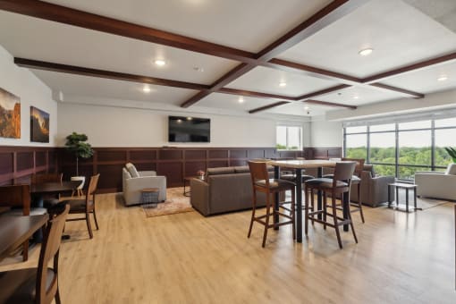 A spacious community room with modern seating arrangements, including high-top tables with chairs and lounge areas with sofas. Large windows offer a scenic view, and a flat-screen TV is mounted on the wall. The room has a bright, inviting atmosphere.