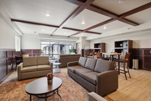 A stylish community lounge with comfortable seating, including sofas and armchairs, a coffee table, and high-top tables with chairs. The space has modern decor, wood accents, and large windows letting in natural light.