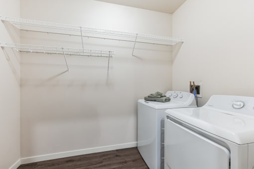 A laundry room featuring a washer and dryer, with wire shelving above for storage. The space has wood-style flooring and a simple, clean design.