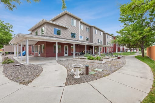 Three-story apartment building with beige and red siding, featuring a covered patio area with outdoor seating. The landscaped courtyard includes a walking path, decorative garden features, and raised garden beds, surrounded by lush greenery and mature trees