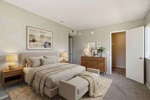 A bedroom with a large bed, a nightstand with a lamp, and a dresser with a plant on top.