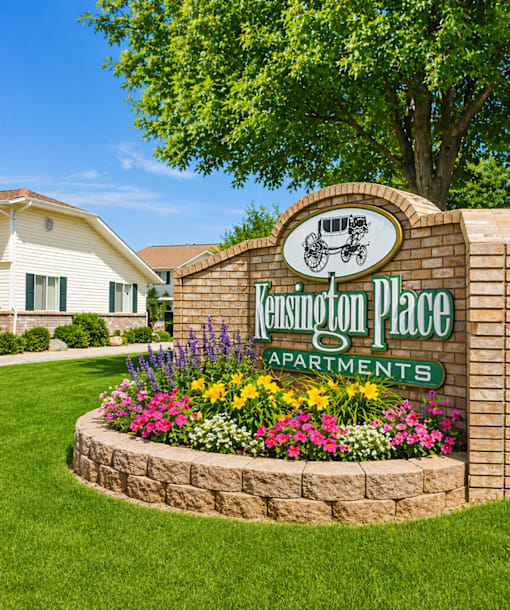 A sign for Kensington Place Apartments is surrounded by flowers and greenery.