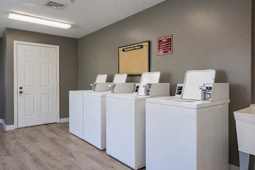Three white washing machines are lined up in a laundry room at Arbors at Hickory Creek Apartments , University Park