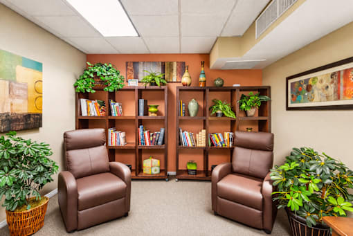 A room with two brown leather chairs, a bookshelf, and potted plants.