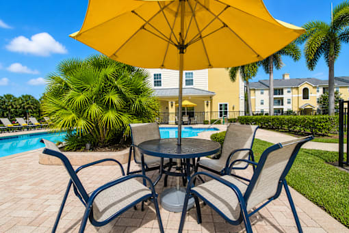 A table with chairs and an umbrella is in the foreground of a pool area.