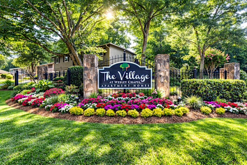 A sign for The Village Apartment Homes is surrounded by flowers and greenery.