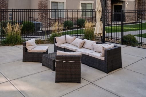 Outdoor lounge area in an apartment community featuring a wicker sectional sofa with beige cushions, a matching armchair, and a glass-top coffee table. The space is set on a paved patio with decorative grasses, a closed patio umbrella, and a black metal fence enclosing the area