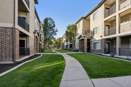 Modern apartment community with three-story buildings featuring brick and stucco exteriors. A winding concrete walkway leads through well-manicured green lawns and mature trees, creating a peaceful residential setting