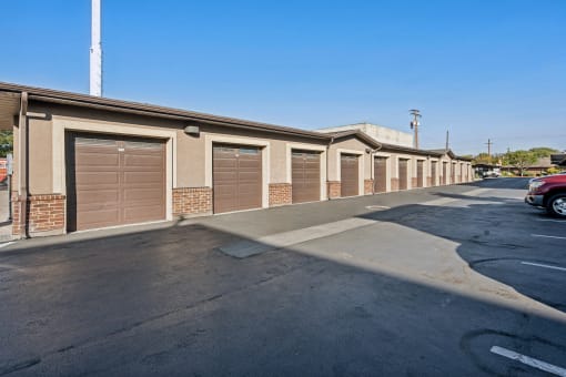 Row of private garage units in an apartment community, featuring brown roll-up doors, beige stucco walls, and brick accents. The area is paved with asphalt and offers secure, enclosed parking for residents