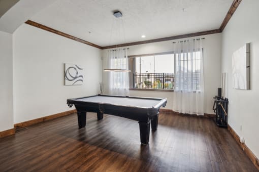Modern apartment clubhouse game room featuring a black pool table, wooden flooring, and large windows with sheer curtains. The space includes a sleek overhead light fixture, abstract wall art, and a cue rack for pool accessories