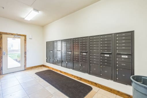 Apartment community mailroom featuring a wall of black metal mailboxes, a tiled floor with a dark entry mat, and a glass door leading outside. The well-lit space provides residents with secure and organized mail storage