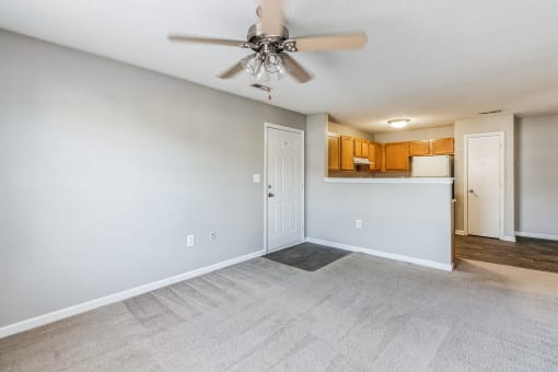 Empty living room with light gray walls and carpeted flooring, featuring a ceiling fan with wooden blades. The room opens into a kitchen with wooden cabinets, white appliances, and a breakfast bar. A white door leads to another area of the home, and there is a mix of carpet and hardwood flooring
