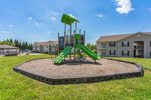 Outdoor playground with green slides, climbing features, and a shaded platform, surrounded by a mulch-filled play area with a black border. The playground is situated on a grassy lawn near multi-story apartment buildings, with a clear blue sky overhead. A swimming pool area with lounge chairs is visible in the background