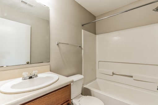 Bathroom with a white bathtub and shower combination, featuring a built-in grab bar and a shower rod above. A vanity with a white sink, wooden cabinet, and beige countertop is next to a toilet. A large mirror is mounted above the sink, reflecting a white door. The walls are painted in a neutral tone, and a towel rack is installed on the side wall