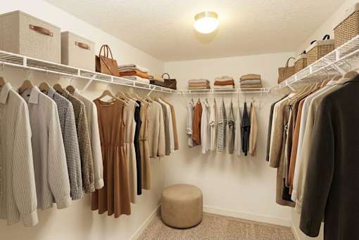 A well organized walk in closet with clothes hanging on shelves and a round ottoman.