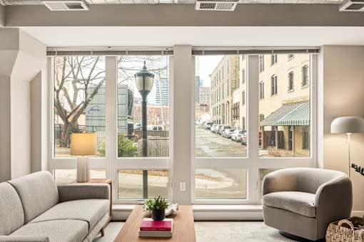 A living room with a grey couch, a grey armchair, a wooden table with a red book and a plant on it, and a view of a street through the windows.