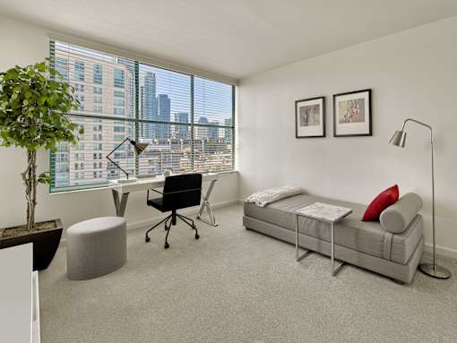 A room with a grey couch, a desk with a chair, a potted plant, and two framed pictures on the wall.