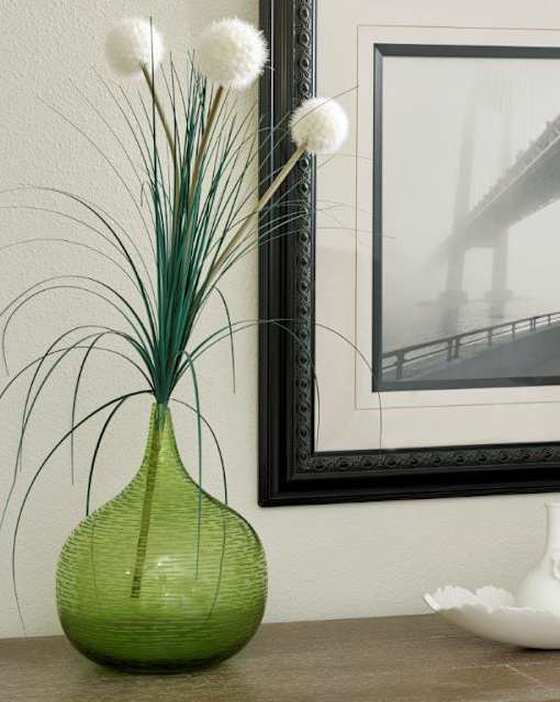 A green vase with white pom poms and long thin stems sits on a table.