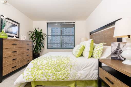A bedroom with a bed, nightstand, and a window with a view of a building.